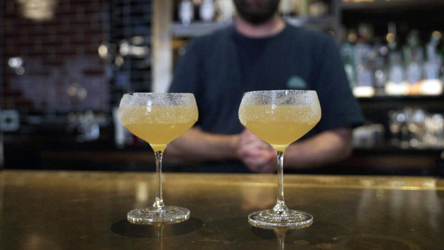 Hawksmoor Chicago beverage director Liam Davy created a twist on the Sidecar while Geoffrey Baer filmed an interview for "Riding the Rails." Two yellow cocktails in coupe glasses sit atop a bar while Liam Davy stands behind the bar out of focus